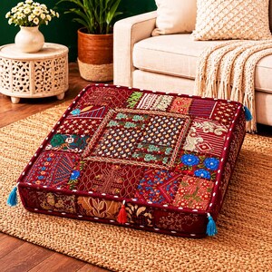 May include: A square, patchwork floor cushion in shades of burgundy, red, and gold. The cushion features a variety of embroidered patterns and textures, with blue and orange tassels at the corners. It is placed on a woven jute rug.