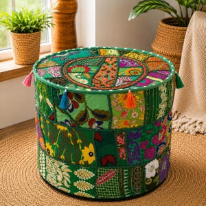 Puede incluir: Un puf cilíndrico redondo con un diseño de retazos de paneles de tela verde con patrones florales y geométricos bordados. La parte superior y los lados están decorados con pequeñas borlas de tela coloridas en rosa, azul y naranja.