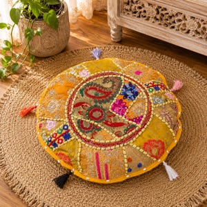 Puede incluir: Cojín de suelo redondo de patchwork en tonos amarillos, naranjas y rojos. El cojín presenta un diseño central de cachemira y está adornado con borlas de colores en los bordes. Se encuentra sobre una alfombra de yute tejida, de unos 70 cm de diámetro.
