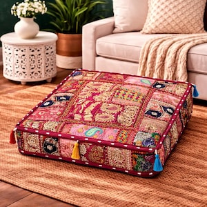 May include: Square floor cushion with a patchwork design in shades of pink, red, and gold. The cushion features intricate embroidery and tassels on the corners. The cushion is placed on a woven rug.