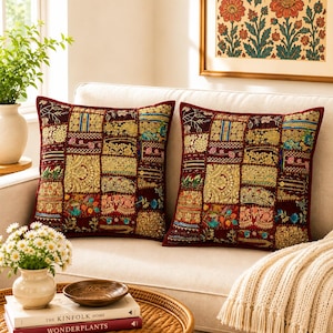 May include: Two square throw pillows featuring a dark maroon base with a patchwork design of embroidered gold, blue, green, and pink floral and geometric patterns. The pillows are decorated with intricate metallic thread work and bead accents.