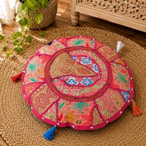 May include: Round, patchwork floor cushion in shades of pink, coral, and gold. The cushion features a floral and paisley pattern with decorative tassels in various colors. The cushion is placed on a round, woven jute rug.