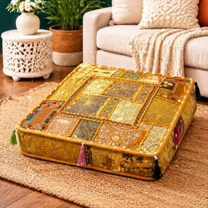 May include: Square floor cushion with a patchwork design in shades of gold, brown, and olive green. The cushion has a decorative border with small white beads and tassels on the corners. It rests on a woven jute rug.