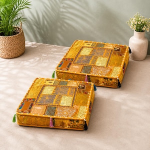 May include: Two square floor cushions featuring a vibrant yellow patchwork design with intricate embroidery, metallic thread accents, and small colorful tassels attached to the corners. The cushions have a low profile and a structured, flat shape.