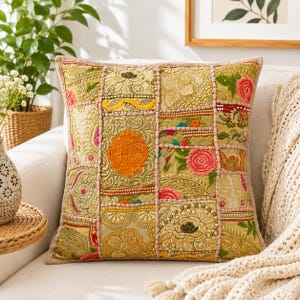 May include: A square decorative cushion featuring a patchwork design of gold, yellow, and olive green fabric panels. The surface is heavily embroidered with floral motifs, intricate beadwork, and sequins in shades of pink, orange, and green.