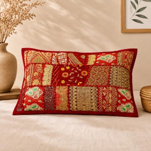 May include: A rectangular throw pillow featuring a patchwork design of red fabric panels. The surface is decorated with intricate gold embroidery, metallic thread accents, small yellow floral beadwork, and green paisley-shaped fabric appliques.