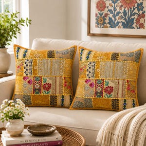 May include: Two square throw pillows featuring a patchwork design of mustard yellow, gold, and beige fabric panels. The pillows are decorated with intricate embroidery, floral motifs, and metallic thread accents in shades of pink, blue, and green.