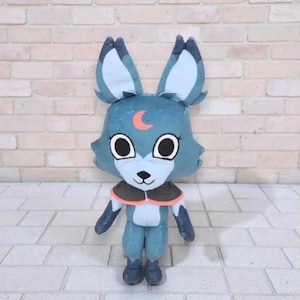 Könnte beinhalten: Ein blaues Plüschfuchs-Spielzeug mit großen Ohren und einem Halbmond auf der Stirn. Der Fuchs hat große, runde Augen und eine kleine, schwarze Nase. Er hat einen braunen und orangefarbenen Kragen. Die Plüschfigur ist etwa 25 cm groß.