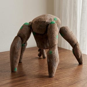 Puede incluir: Araña de peluche marrón con seis patas articuladas y un cuerpo redondeado. El juguete tiene detalles verdes en las patas y el cuerpo. La araña está hecha de un material suave y texturizado.