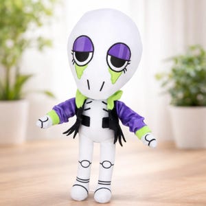 Puede incluir: Una muñeca de peluche con cabeza y cuerpo blancos, con sombra de ojos morada, ojos negros y lágrimas verdes. Viste una chaqueta morada con chaleco negro y cuello verde. La muñeca tiene brazos y piernas blancos con detalles negros, de pie sobre una superficie de madera.