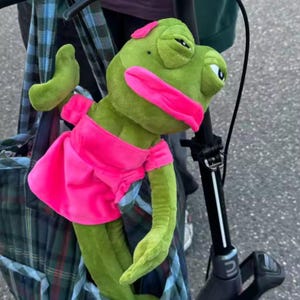 Könnte beinhalten: Eine grüne Froschpuppe mit einem pinkfarbenen Kleid und Stirnband. Die Puppe hat große rosa Lippen und posiert mit einem erhobenen Arm. Die Puppe ist an einem schwarzen Roller befestigt.