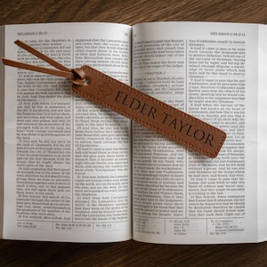 May include: An open book with a brown leather bookmark. The bookmark has the text "ELDER TAYLOR" and a Texas outline. The book is on a wooden surface.