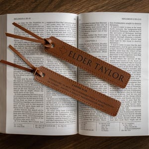 May include: Two brown leather bookmarks with text, resting on an open book. One bookmark reads "ELDER TAYLOR" and features a Texas shape. The other bookmark has text from 2 Nephi 31:20. The book is open on a wooden surface.