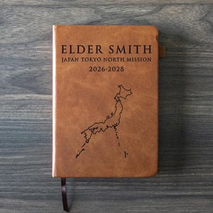 Puede incluir: Un diario de cuero marrón con el texto "ELDER SMITH JAPAN TOKYO NORTH MISSION 2026-2028" y un mapa de Japón. El diario tiene una cinta de marcador.