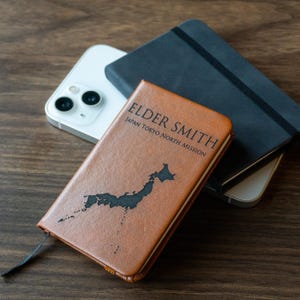 May include: A brown leather journal with the text "ELDER SMITH JAPAN TOKYO NORTH MISSION" and a black map of Japan. A black notebook and a white smartphone are also in the image.
