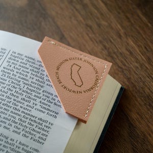 May include: A peach-colored leather bookmark with white stitching, placed on the corner of a book. The bookmark features a circular design with the text "Newport Beach Mission Sister Johnson California" and a California state outline.