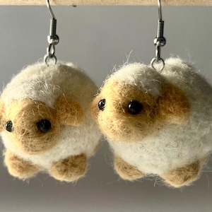 Another picture of the earrings hanging. A pair of needle-felted sheep earrings. Each earring has a fuzzy, textured ball shape with small legs, heads and tails. The earrings hang from silver-colored fish hook ear wires.