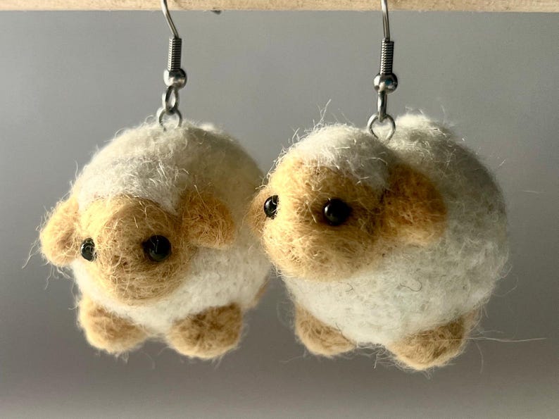 Another picture of the earrings hanging. A pair of needle-felted sheep earrings. Each earring has a fuzzy, textured ball shape with small legs, heads and tails. The earrings hang from silver-colored fish hook ear wires.