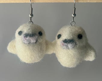 以下が含まれることがあります： オフホワイトのアザラシのイヤリングのペア。それぞれのアザラシは黒い目とピンクとグレーの口をしています。イヤリングは柔らかくふわふわした素材でできており、銀色のフックから吊り下げられています。
