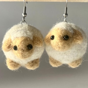 A pair of needle-felted sheep earrings. Each earring has a fuzzy, textured ball shape with small legs, heads and tails. The earrings hang from silver-colored fish hook ear wires.