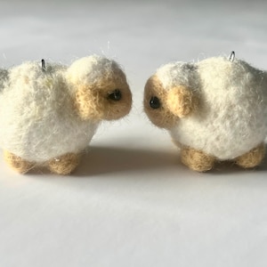 Another picture of the earrings facing each other. A pair of needle-felted sheep earrings. Each earring has a fuzzy, textured ball shape with small legs, heads and tails. The earrings hang from silver-colored fish hook ear wires.