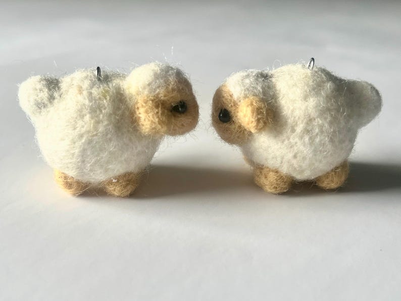 Another picture of the earrings facing each other. A pair of needle-felted sheep earrings. Each earring has a fuzzy, textured ball shape with small legs, heads and tails. The earrings hang from silver-colored fish hook ear wires.