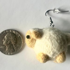 Another picture of the earring. It is shown next to a US quarter for size comparison.  Needle-felted sheep earring. It has a fuzzy, textured ball shape with small legs, head and tail. The earring has a silver-colored fish hook earring wire.