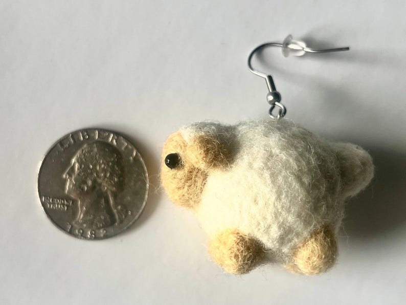 Another picture of the earring. It is shown next to a US quarter for size comparison.  Needle-felted sheep earring. It has a fuzzy, textured ball shape with small legs, head and tail. The earring has a silver-colored fish hook earring wire.