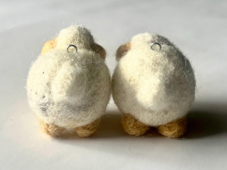 Another picture of the earrings from the back. A pair of needle-felted sheep earrings. Each earring has a fuzzy, textured ball shape with small legs, heads and tails. The earrings hang from silver-colored fish hook ear wires.
