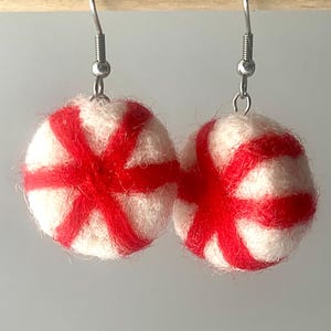 Puede incluir: Un par de pendientes redondos de lana fieltrada con una base blanca y rayas rojas vibrantes que irradian desde el centro. Cada esfera está unida a un gancho de metal plateado, creando un diseño colgante que se asemeja a un caramelo de menta.