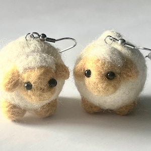 Another picture of the earrings standing in place. A pair of needle-felted sheep earrings. Each earring has a fuzzy, textured ball shape with small legs, heads and tails. The earrings hang from silver-colored fish hook ear wires.