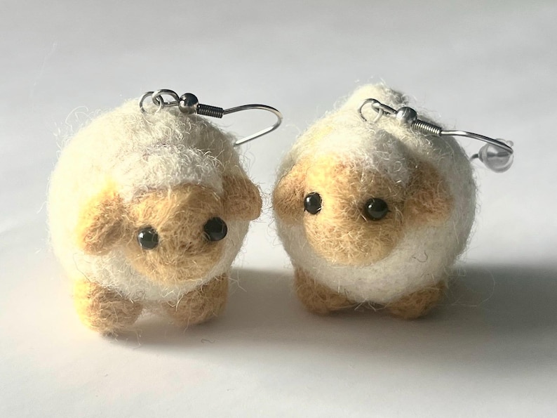 Another picture of the earrings standing in place. A pair of needle-felted sheep earrings. Each earring has a fuzzy, textured ball shape with small legs, heads and tails. The earrings hang from silver-colored fish hook ear wires.