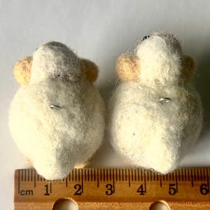 Another picture of the earrings. They are shown next to a ruler and are approximately 2.5 centimeters in width.  Needle-felted sheep earrings. They have uzzy, textured ball shapes with small legs, heads and tails. The earring has a silver-colored fish hook earring wire.
