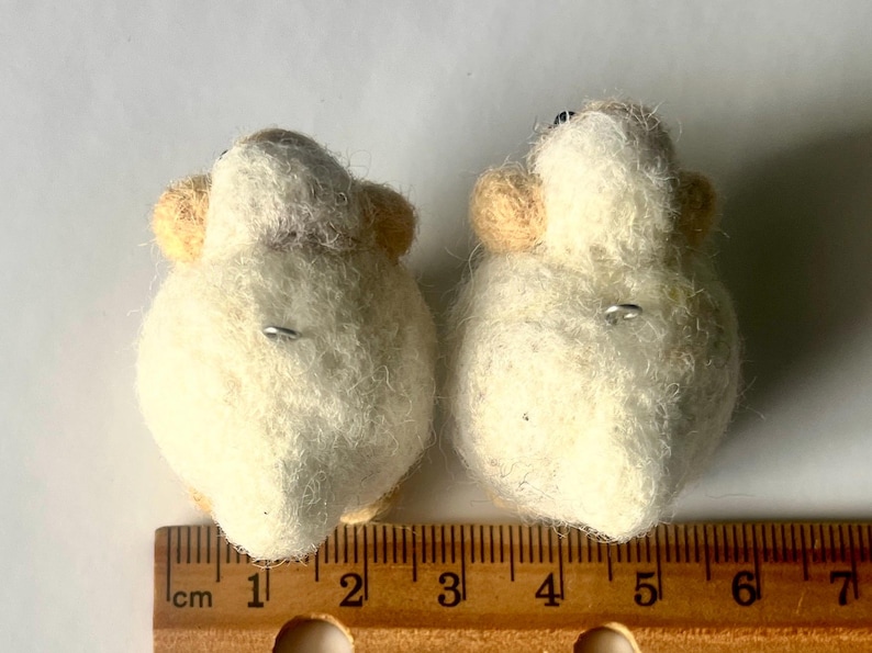 Another picture of the earrings. They are shown next to a ruler and are approximately 2.5 centimeters in width.  Needle-felted sheep earrings. They have uzzy, textured ball shapes with small legs, heads and tails. The earring has a silver-colored fish hook earring wire.
