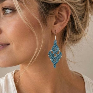 Könnte beinhalten: Ein hängender Ohrring aus blauem Makramee-Garn, das in einem rautenförmigen Muster geknotet ist. Das Design enthält kleine silberfarbene Metallperlen, die in die geflochtene Struktur eingearbeitet sind und an einem silberfarbenen Metallhaken hängen.