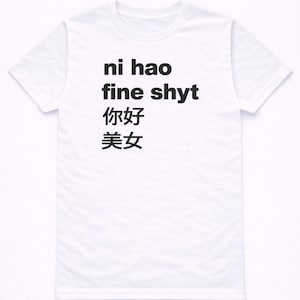 Könnte beinhalten: Weißes T-Shirt mit schwarzem Text, der "ni hao fine shyt" auf Englisch und in chinesischen Schriftzeichen lautet. Das Shirt hat einen Rundhalsausschnitt und scheint aus Baumwolle zu sein.