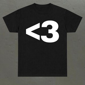 Könnte beinhalten: Ein schwarzes T-Shirt mit kurzen Ärmeln und einem großen, weißen Grafikdruck auf der Brust. Die Grafik besteht aus den Zeichen < und 3, die zusammen ein Herzsymbol bilden. Das T-Shirt hat einen klassischen Rundhalsausschnitt und einen geraden Saum.