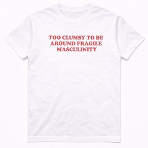 Könnte beinhalten: Ein weißes T-Shirt mit kurzen Ärmeln und dem Text "TOO CLUMSY TO BE AROUND FRAGILE MASCULINITY", der in roten Großbuchstaben auf der Brustmitte aufgedruckt ist. Das Kleidungsstück hat einen Rundhalsausschnitt und eine Standardpassform.
