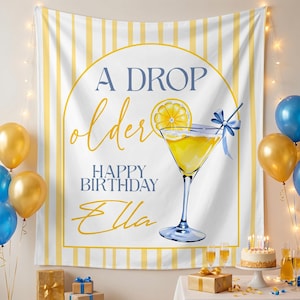 May include: A white and yellow striped backdrop with the text "A Drop Older Happy Birthday Ella" and a watercolor illustration of a cocktail. Gold and blue balloons and a birthday cake are also visible.