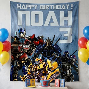 Peut inclure: Scène de fête d'anniversaire avec une bannière « JOYEUX ANNIVERSAIRE ! » et un nom, plus le chiffre « 3 ». La bannière a un fond bleu et des images de personnages Transformers. Des ballons bleus, rouges et jaunes sont visibles.