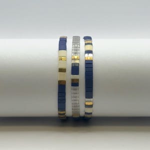 May include: Three beaded bracelets with a minimalist design. The bracelets feature a combination of navy blue, cream, silver, and gold-colored beads. The beads are flat and rectangular, creating a modern, geometric look. The bracelets are displayed against a white background.