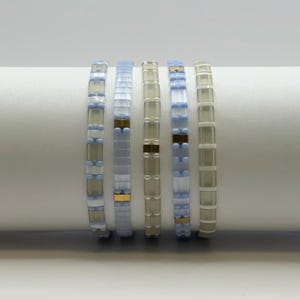 May include: Six beaded bracelets in shades of blue, white, and clear. The bracelets have a rectangular bead design with gold-colored accents. The bracelets are displayed on a white surface.
