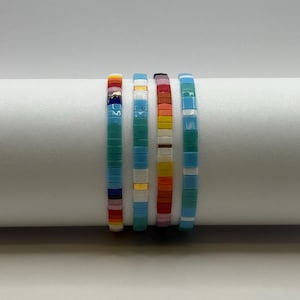May include: Four colorful beaded bracelets. Each bracelet features a different color scheme, including turquoise, blue, white, yellow, orange, and red. The bracelets are made of small, flat beads strung together, creating a vibrant and eye-catching accessory.