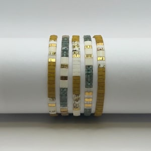 May include: Six beaded bracelets in shades of olive green, white, and gold. The bracelets are made of small, rectangular beads strung together. The design includes gold-colored accents and a mix of solid and patterned beads. The bracelets are stacked together.