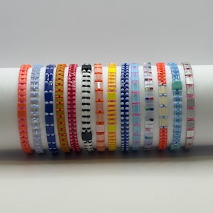 May include: A collection of colorful beaded bracelets in various shades, including orange, blue, yellow, and pink. The bracelets are displayed on a white cylindrical surface, showcasing their diverse designs and patterns. Each bracelet is approximately 0.5 inches wide.