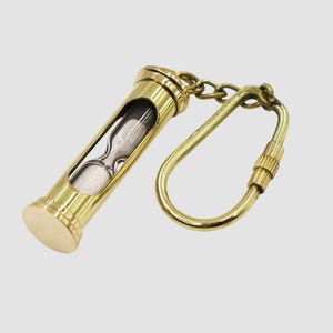 May include: A brass hourglass keychain with a clear glass center containing white sand. The keychain has a gold-colored clasp and chain. The hourglass is a classic design, perfect for a vintage or steampunk aesthetic.