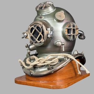 Puede incluir: Un casco de buceo vintage con acabado metálico, ventanas de visión redondas y detalles intrincados. El casco descansa sobre una base de madera, con una cuerda enrollada que añade a su estética náutica.