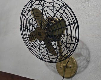 May include: A wall-mounted electric fan featuring a black wire cage and metallic gold-toned blades. The center of the fan displays a circular emblem with the text "MADE FOR ROYAL NAVY LONDON". The base is a circular gold-toned plate attached to the wall.