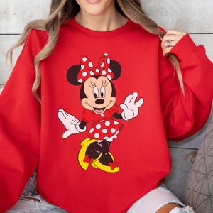Op de afbeelding: Een rode sweatshirt met een Minnie Mouse-afbeelding. De afbeelding toont Minnie Mouse in een rode jurk met witte stippen, een bijpassende strik en gele schoenen. De sweatshirt heeft een effen rode kleur.