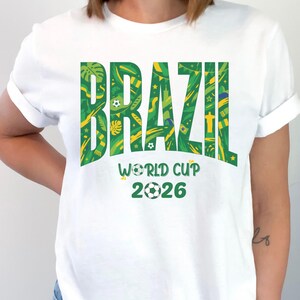 May include: White t-shirt with the word "BRAZIL" in large green letters filled with a tropical pattern. Below, the text "WORLD CUP 2026" is displayed with soccer ball graphics, celebrating the upcoming tournament.